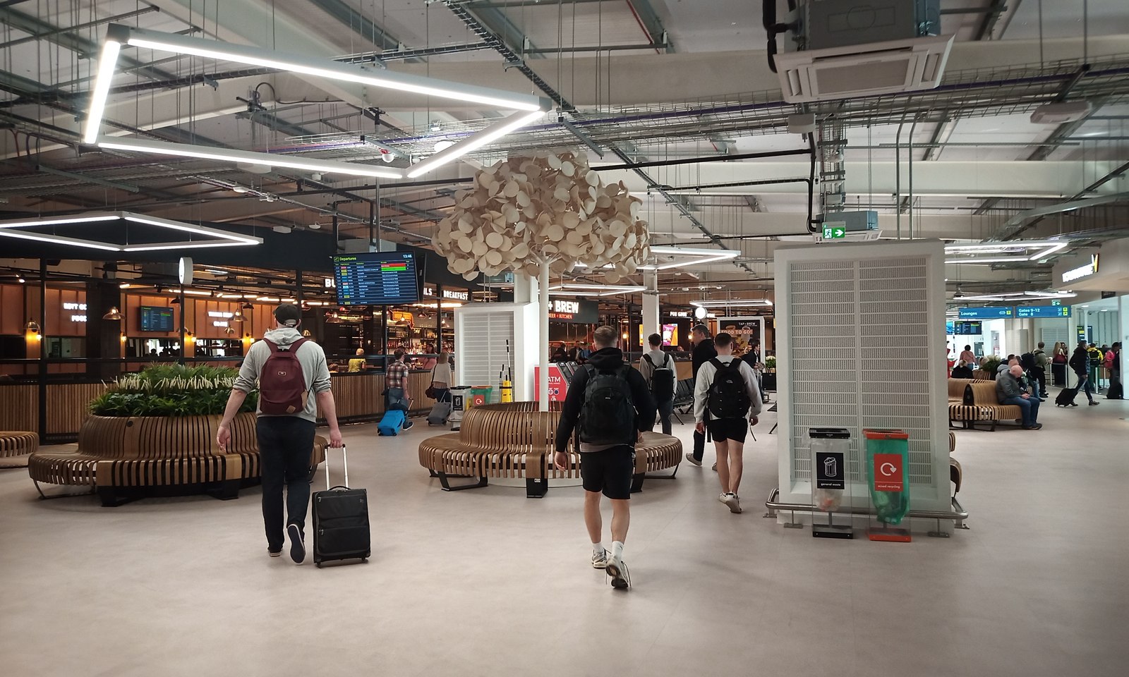 Passengers in the terminal departure lounge, with flight information screens, seating and overhead cable trays.